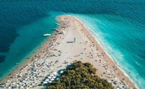 Luftaufnahme eines Strandes mit türkisfarbenem Wasser auf beiden Seiten, der eine schmale Landzunge bildet. Mehrere Menschen sonnen sich, schwimmen und entspannen unter Sonnenschirmen. Boote liegen in Ufernähe vor Anker und am Fuße des Strandes erstreckt sich dichtes Grün.