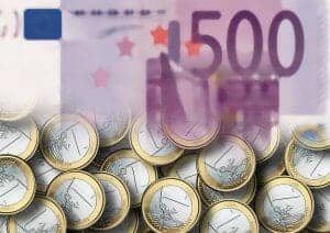 Im Hintergrund ist eine große, teilweise verblasste 500-Euro-Banknote zu sehen, im Vordergrund ein Stapel 1-Euro-Münzen. Das Bild zeigt den Kontrast zwischen dem höheren Nennwert der Banknoten und den kleineren Nennwerten der Münzen.