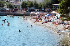 Ein überfüllter Strand mit vielen Menschen, die im klaren blauen Wasser schwimmen und sich am felsigen Ufer sonnen. Sonnenschirme und Handtücher sind entlang des Strandes ausgebreitet, im Hintergrund sind Bäume und Gebäude zu sehen.