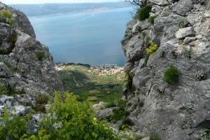 Durch eine Lücke zwischen felsigen Klippen ist ein Blick auf ein Küstendorf zu sehen. Im Vordergrund sind schroffe, graue Felsen mit einigen grünen Pflanzen und gelben Blumen zu sehen, darunter eine üppige Landschaft und im Hintergrund ein Gewässer.