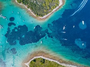 Luftaufnahme eines Küstengebiets mit klarem türkisfarbenem Wasser und vielen kleinen Booten, die überall verstreut sind. Die Küste zeichnet sich durch üppige grüne Vegetation, felsige Ufer und Sandstrände aus. Das Meer weist verschiedene Blautöne auf, die auf Tiefenunterschiede hinweisen.