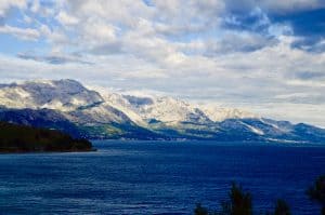 Eine ruhige Landschaft mit einer riesigen, blauen Wasserfläche im Vordergrund und einer Bergkette im Hintergrund unter einem teilweise bewölkten Himmel. Die Berge sind in Sonnenlicht getaucht, das die Bergrücken und Hänge hervorhebt.