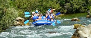 Eine Gruppe von Menschen mit Helmen und Schwimmwesten paddelt auf einem blauen Floß durch einen Fluss mit Wildwasser und Felsen. Die Umgebung ist üppig und grün mit verschiedenen Vegetationsarten. Eine Person im hinteren Teil des Floßes scheint das Team zu führen.