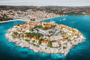 Luftaufnahme einer malerischen Küstenstadt mit Gebäuden mit roten Dächern, die sich auf einer felsigen Halbinsel umgeben von türkisfarbenem Wasser befinden. Im Hintergrund ist das Festland mit weiteren Gebäuden zu sehen, in der Bucht liegen Segelboote vor Anker.