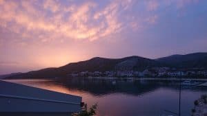 Ein ruhiger Blick auf einen Sonnenuntergang über einer ruhigen Uferpromenade mit Bergen im Hintergrund. Der Himmel ist in Rosa- und Lilatönen bemalt, die sich im ruhigen Wasser darunter spiegeln. Entlang der Küste sind Häuser und Boote zu sehen.