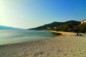 Ein ruhiger Strand mit goldenem Sand und ruhigem, klarem Wasser unter einem strahlend blauen Himmel. Vereinzelte Menschen faulenzen unter ein paar Sonnenschirmen am Ufer. In der Ferne sind grüne Hügel und ein paar verstreute Häuser zu sehen.