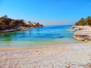 Eine ruhige Strandszene mit klarem, blauem Wasser, Booten, die in Ufernähe schwimmen, und ein paar Gebäuden, die auf der linken Seite zwischen Bäumen eingebettet sind. Der Sandstrand geht in eine felsige Küste über und der helle, wolkenlose Himmel verstärkt die ruhige Atmosphäre.