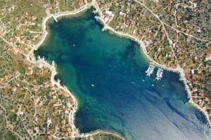 Eine Luftaufnahme einer Küstenstadt mit Gebäuden mit roten Dächern, die eine blaue, unregelmäßig geformte Bucht umgeben. Die Stadt ist dicht am Wasser besiedelt und zwischen den Gebäuden sind Grünflächen verteilt. An den Rändern der Bucht liegen Boote vor Anker.