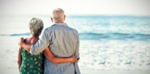 Ein älteres Paar steht an einem Strand mit Blick auf das Meer. Der Mann hat seinen Arm um die Schultern der Frau gelegt und sie hat ihren Arm um seine Taille gelegt. Sie blicken beide auf das ruhige Meer, auf dem an diesem hellen, klaren Tag sanfte Wellen zu sehen sind.