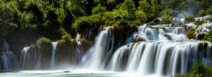 Ein üppiger, grüner Wald umgibt eine Reihe von Wasserfällen, wobei das Wasser in mehreren Stufen sanft über Felsen in einen ruhigen, türkisfarbenen Teich darunter fließt. Das durch die Bäume einfallende Sonnenlicht verstärkt das kräftige Grün und schafft eine sanfte, neblige Atmosphäre.