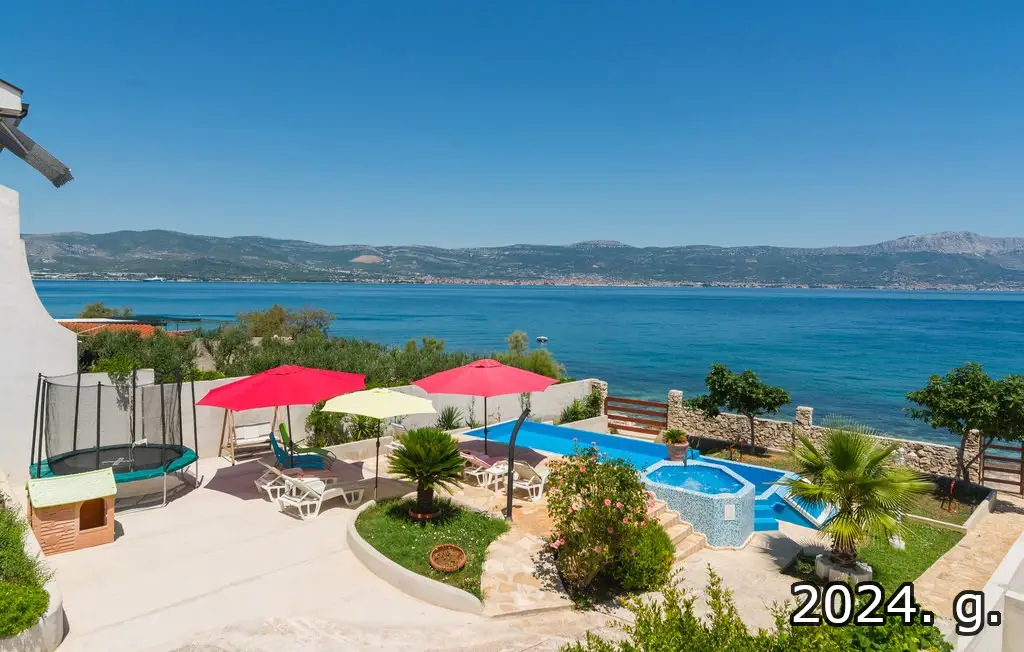 Eine malerische Küstenansicht in Kroatien bietet eine Terrasse mit einem Trampolin, Sonnenliegen unter bunten Sonnenschirmen, einen kleinen Pool und üppiges Grün. Das weite Meer und die fernen Berge bilden den Hintergrund, darüber ein klarer blauer Himmel. Der Text „2024. g.“ erscheint in der unteren rechten Ecke – Immobilien Kroatien in der Tat!