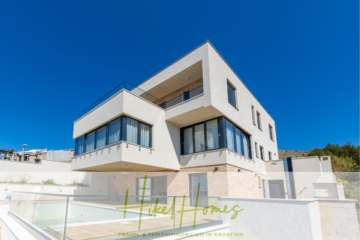 Ein modernes, weißes, würfelförmiges Haus mit großen Fenstern, einem Balkon und einem klaren, blauen Himmel. Die untere Ebene verfügt über einen Pool und die umliegende Landschaft ist teilweise sichtbar. - Ein Paradies mit atemberaubendem Meerblick! 4 SZ, Pool, Solar, ...