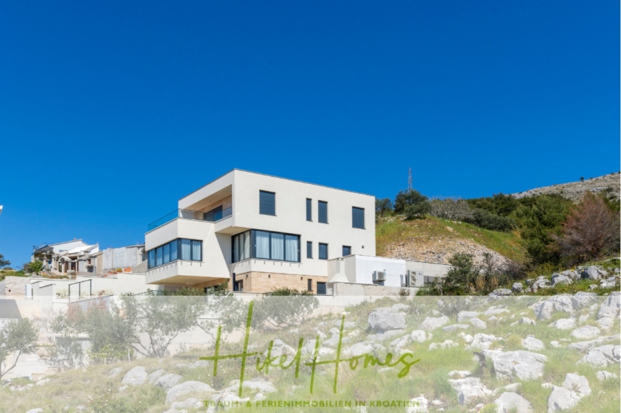 Ein modernes, weißes, kastenförmiges Haus liegt auf einem felsigen Hügel unter einem klaren blauen Himmel. Das Haus verfügt über große Fenster und Balkone. Das Haus ist von felsigem Gelände und spärlichem Grün umgeben. Am unteren Bildrand befinden sich ein Logo und eine Textüberlagerung. - Ein Paradies mit atemberaubendem Meerblick! 4 SZ, Pool, Solar, ...