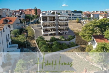 Ein modernes mehrstöckiges Gebäude mit großen Balkonen liegt auf einem Hügel, umgeben von Grün und Nachbarhäusern. Im Vordergrund steht der Text „Hikel Homes Traum- & Ferienimmobilien in Kroatien“. - Luxus Meerblick 77m² Ensemble mit 2 Schlafzimmern, Terrasse und Südausrichtung (1. OG)