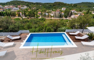 Eine ruhige Szene im Freien mit einem rechteckigen Swimmingpool, umgeben von Holzterrassen und weißen Sonnenliegen. Der Hintergrund zeigt üppiges Grün und einen Blick auf ein kleines Dorf mit Häusern mit roten Dächern auf sanften Hügeln. - 2 Meerblick Villen + freies Grundstück ideal zur Vermietungauf der Insel Dugi Otok jeweils mit Pool