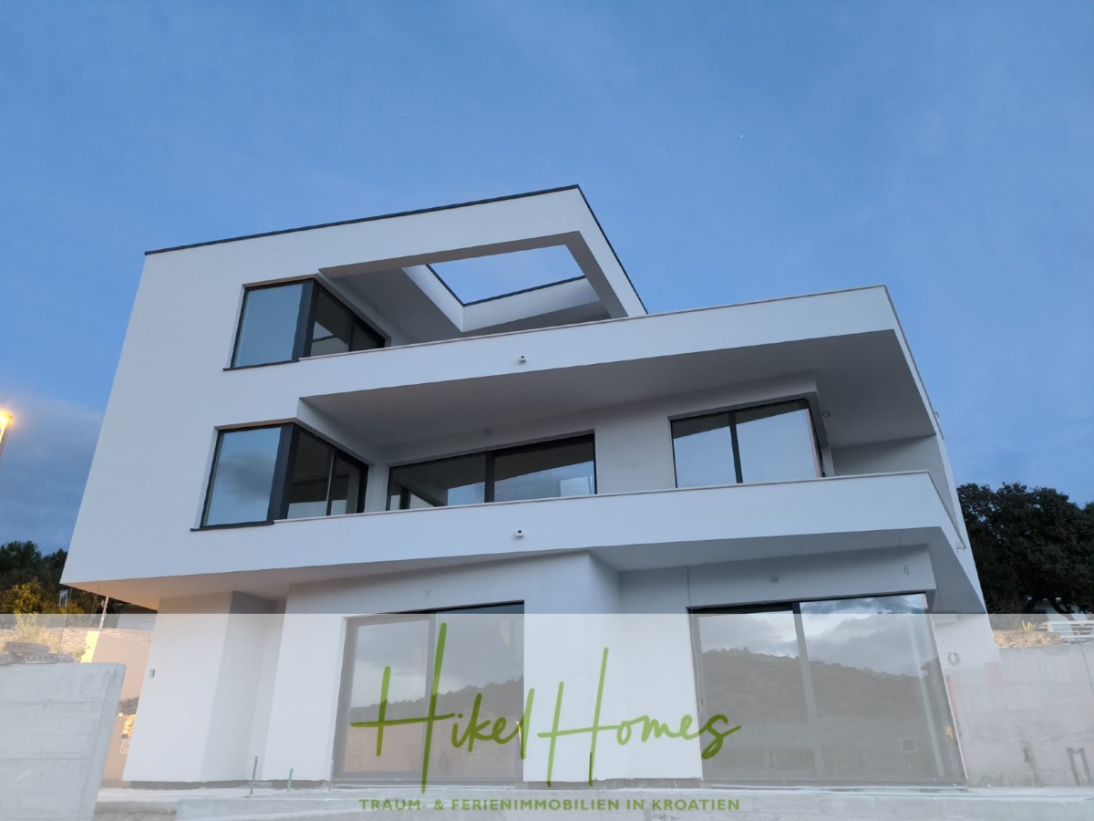 Ein modernes, mehrstöckiges weißes Gebäude mit großen rechteckigen Fenstern und Balkonen. Das Logo „Hikel Homes“ ist unten in Grün überlagert. Der Text darunter lautet: „TRAUM + FERIENIMMOBILIEN IN KROATIEN“. Der Himmel ist klar und die Fassade ist minimalistisch. - Einziehen im Sommer - Meerblick Neubau-Luxusvilla mit Einrichtung, Infinitypool & Raumkonzept