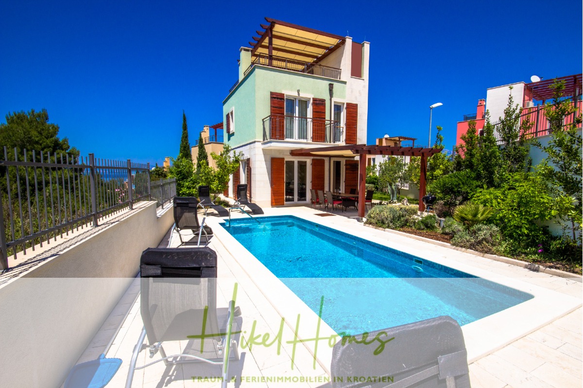 Eine moderne Villa mit grün-roter Außenfassade und einem Pool, der von Liegestühlen umgeben ist. Der Himmel ist klar und blau, mit viel Grün und anderen farbenfrohen Häusern in der Nähe. Die Szene ist hell und sonnig und schafft eine lebendige Urlaubsatmosphäre. - Villa im Mittelmeerstil mit 3 SZ, Pool, Dachterrasse mit Meerblick – möbiliert (15)