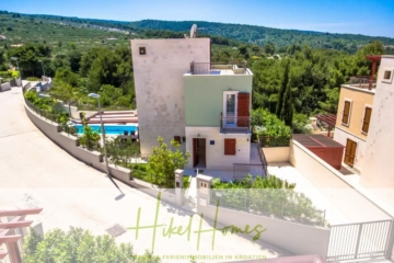 Blick auf ein modernes Haus am Hang mit Balkon, umgeben von üppigem Grün und anderen Häusern. Im Hintergrund ist ein Swimmingpool zu sehen. Der Himmel ist klar und blau und unten steht „Hikel Homes“. - Villa im Mittelmeerstil mit 3 SZ, Pool, Dachterrasse mit Meerblick – möbiliert (15)