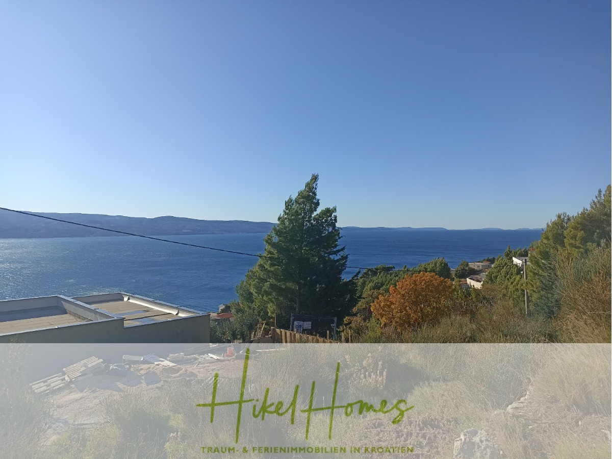 Malerische Aussicht auf eine Küstenlandschaft mit klarem, blauem Himmel. Im Vordergrund sind Bäume und das Dach eines Gebäudes zu sehen. Das Meer erstreckt sich bis zu den Hügeln in der Ferne. Unten befindet sich eine Textüberlagerung mit Branding. - 1515m² Bauland mit fertigen Bauprojekt in Panoramahanglage mit traumhaften Rundumblick aufs Meer