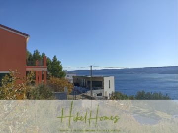 Ein Blick auf eine Küstenregion mit einem terrakottafarbenen Gebäude auf der linken Seite und dem Meer im Hintergrund. Die Gebäude sind von Grün umgeben und der Himmel ist klar und blau. Im Vordergrund sind die Worte „Hikel Homes“ mit einem Slogan auf Deutsch zu sehen. - 1515m² Bauland mit fertigen Bauprojekt in Panoramahanglage mit traumhaften Rundumblick aufs Meer