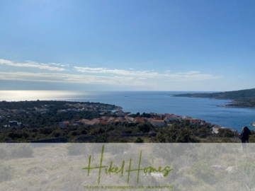 Malerische Aussicht auf ein kroatisches Küstendorf, eingebettet in Grün, mit Blick auf ein ruhiges blaues Meer unter einem hellen, klaren Himmel. Unten ist ein grünes Logo mit dem Text „Hikel Homes“ eingeblendet. - 805m² Bauland in Panoramahanglage mit traumhaften Rundumblick aufs Meer und die vorgelagerten Inseln