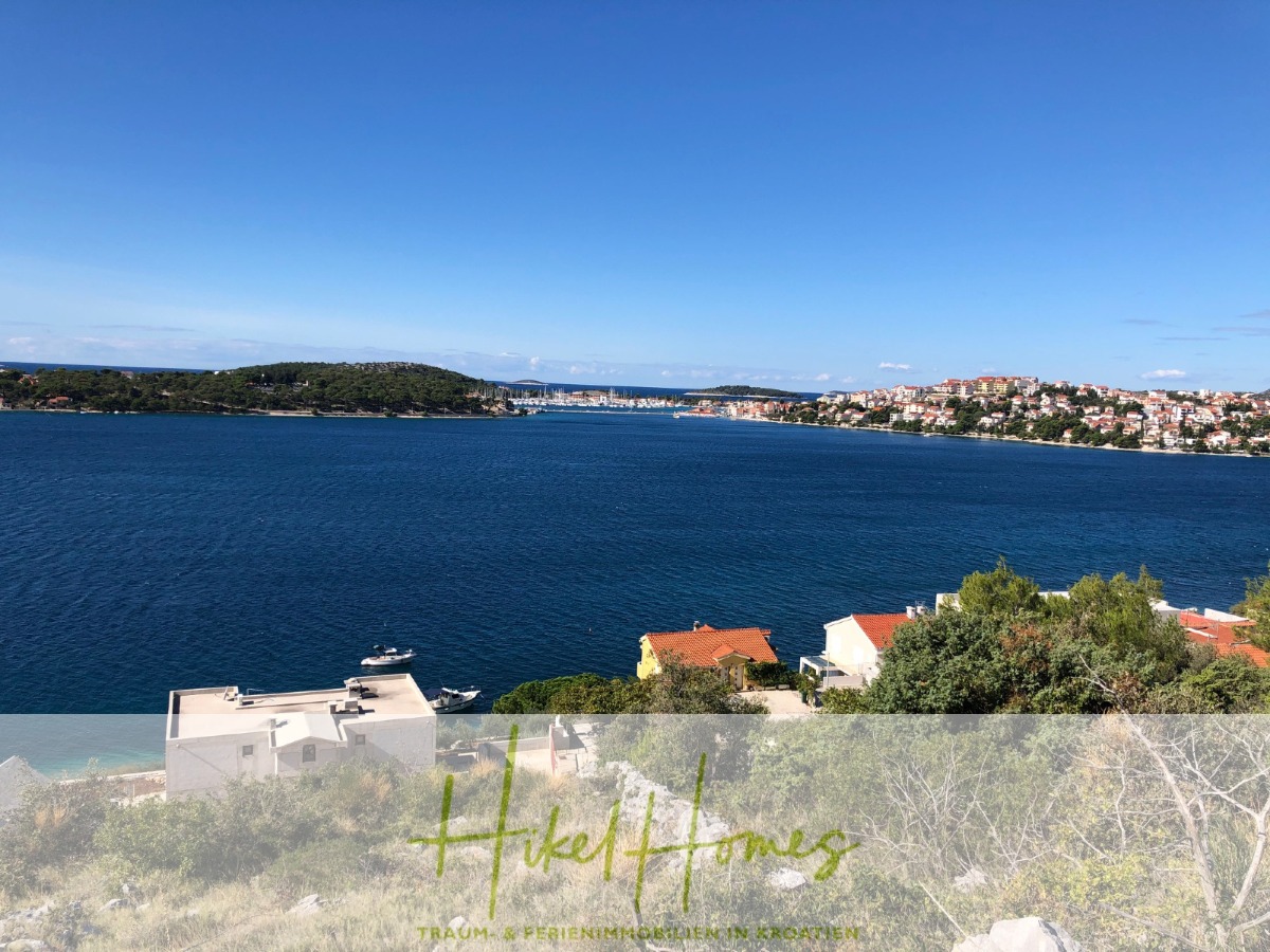 Eine malerische Aussicht auf eine Küstenstadt mit Häusern und üppigem Grün im Vordergrund und Blick auf ein weites blaues Meer. An der entfernten Küste sind weitere Stadtgebäude unter einem klaren blauen Himmel zu sehen.