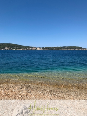 Ein klarer Tag am Strand mit ruhigen blauen Wellen, die sanft gegen eine felsige Küste plätschern. Unter dem strahlend blauen Himmel ist eine entfernte Insel mit üppigem Grün zu sehen. - Einmaliges 2. Reihe mit Meerzugang 1834m² Bauland mit Baugenehmigung für 2 Villen