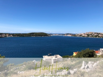 Eine malerische Aussicht auf eine Küstenstadt mit blauem Wasser und grünen Hügeln unter einem klaren blauen Himmel. Im Vordergrund sind ein weißes Gebäude und etwas Vegetation zu sehen. Unten steht in grüner Schrift „Hikel Homes“. - Einmaliges 2. Reihe mit Meerzugang 1834m² Bauland mit Baugenehmigung für 2 Villen