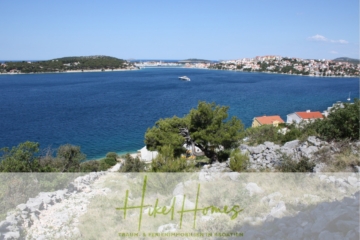 Eine malerische Aussicht auf eine Küstenlandschaft mit einem ruhigen blauen Meer, einem kleinen Boot, felsigen Ufern und Häusern mit roten Dächern. Im Hintergrund sind grüne Hügel und verstreute Gebäude entlang der Küste zu sehen. - Einmaliges 2. Reihe mit Meerzugang 1834m² Bauland mit Baugenehmigung für 2 Villen