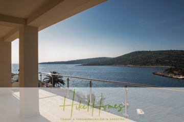 Ein geräumiger Balkon mit Glasgeländer bietet Aussicht auf ein ruhiges Meer und grüne Hügel unter einem klaren blauen Himmel. Im Wasser ist ein kleines Boot zu sehen, und links steht eine Palme. Der Text unten erwähnt „Hikel Homes“ und Ferienimmobilien in Kroatien. - Villa Ana – 250m² WFL in 1. Reihe zum Meer / 4 SZ / Neubau / Sofort bezugsfertig