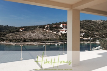 Blick von einem geräumigen, überdachten Balkon auf eine ruhige Bucht mit klarem Wasser und einer grünen, hügeligen Landschaft. An den Hängen sind mehrere Häuser zu sehen. Der Himmel ist klar und im Vordergrund ist ein Glasgeländer zu sehen. - Villa Ana – 250m² WFL in 1. Reihe zum Meer / 4 SZ / Neubau / Sofort bezugsfertig